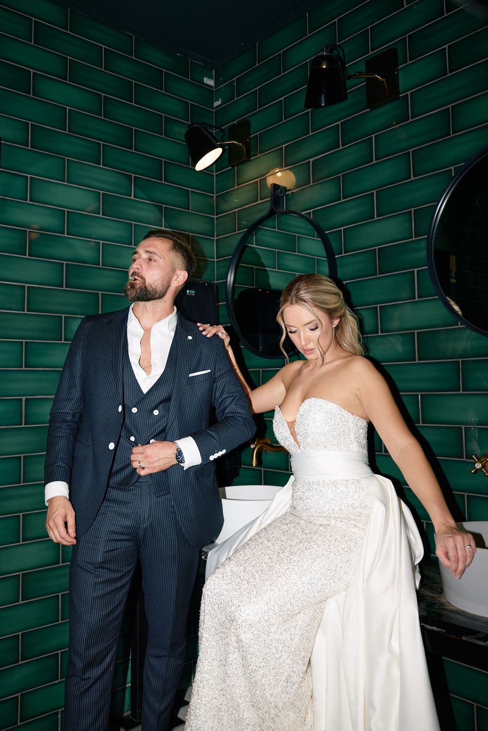 Bride and groom pose elegantly in a green tiled bathroom, she in a strapless gown, he in a navy three-piece suit.
