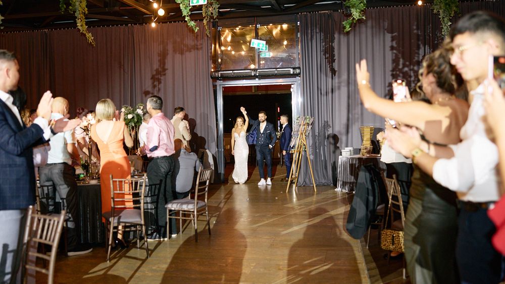 Newlyweds walk through applauding guests at elegant indoor wedding reception with string lights.