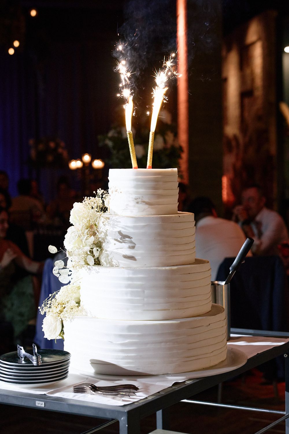 Four-tier white wedding cake with sparklers and white floral decorations at reception.