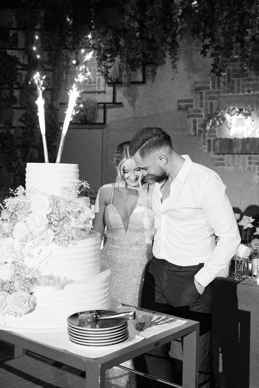 Bride and groom cutting tiered wedding cake with sparklers in black and white photo.