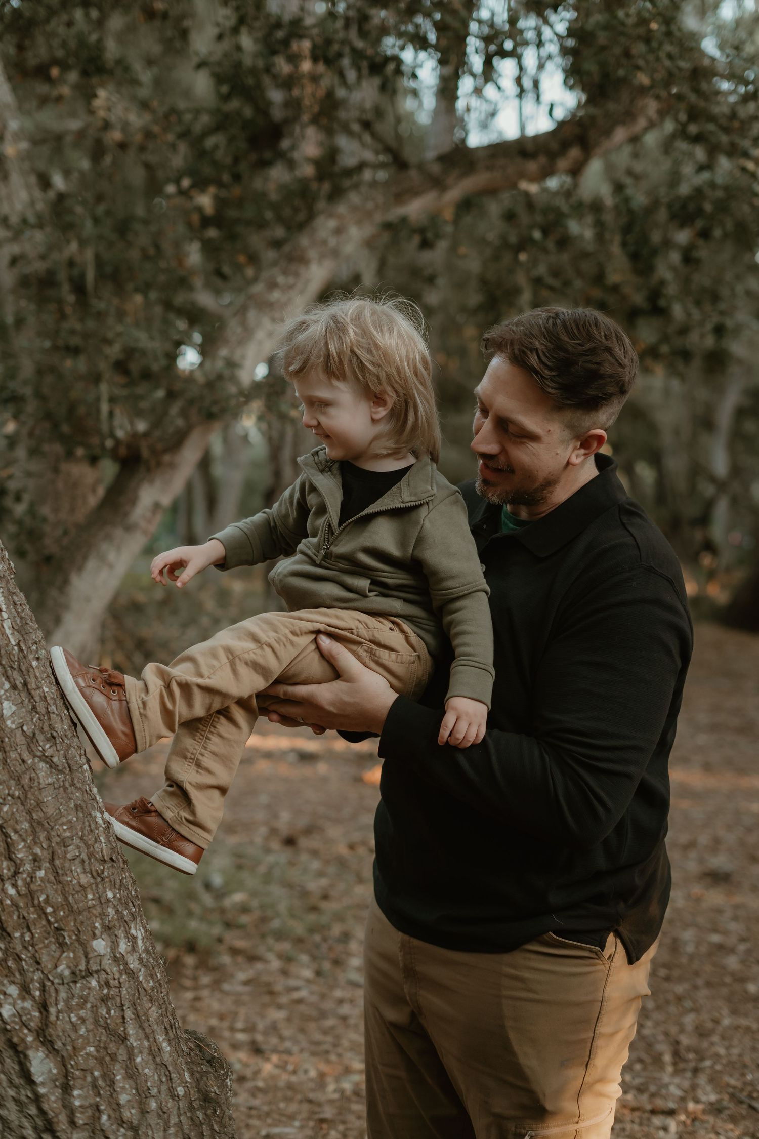 A father holds his young toddler son near a tree trunk in a wooded outdoor setting during a family photo session.