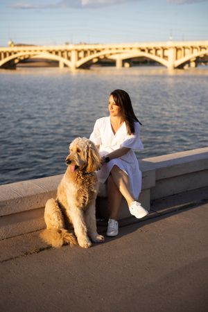 A person in white sits with a fluffy golden-colored dog on a concrete wall by the water during golden hour.