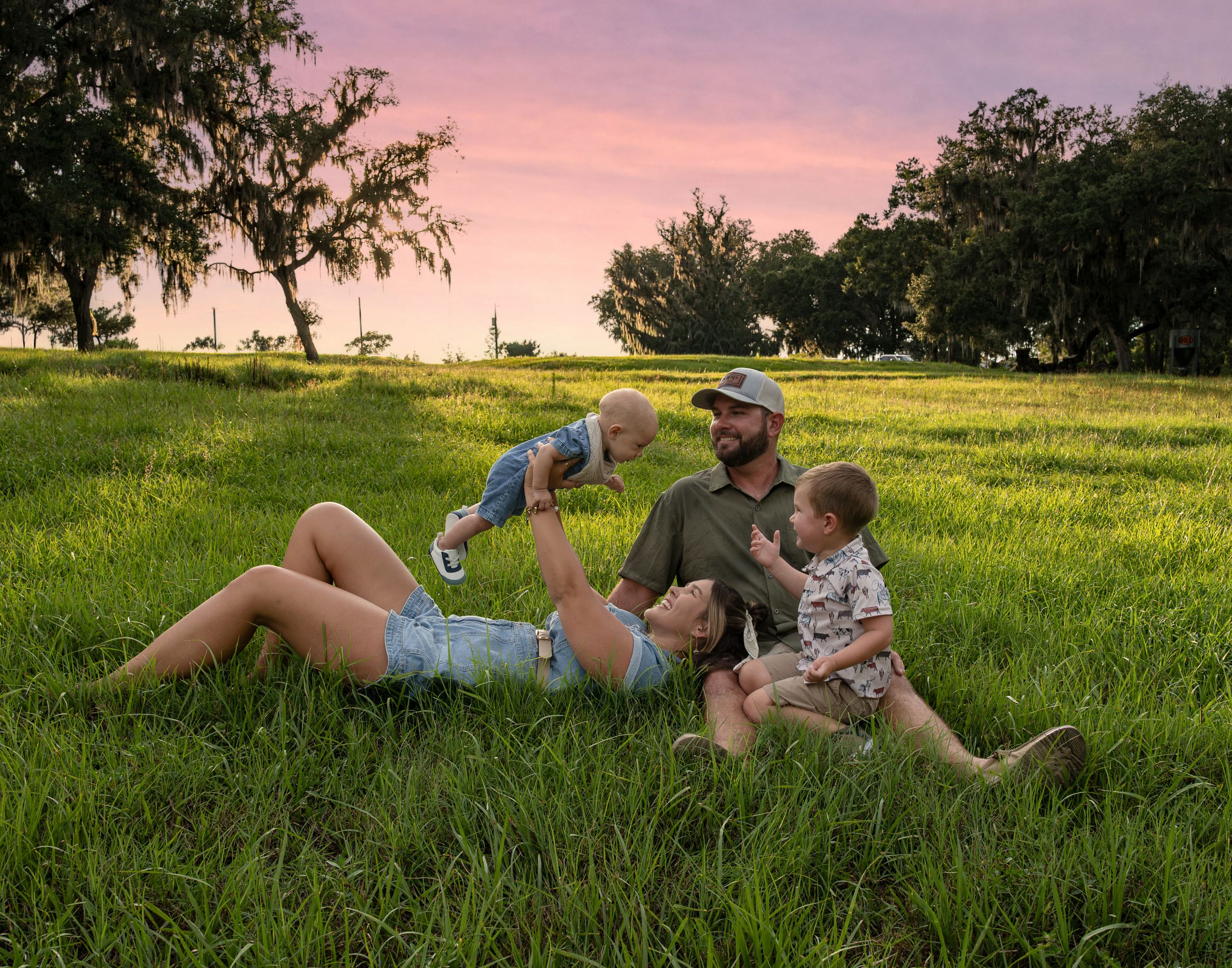 Family of four enjoys a relaxed evening on a lush lawn, lifting a toddler into the air beneath trees and a pastel sunset sky.
