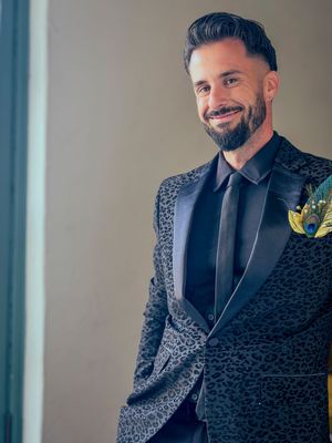 Smiling man in a patterned tuxedo jacket with black tie and floral boutonniere at formal event.