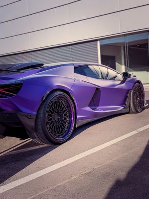 A sleek purple Lamborghini supercar parked on a lot, showcasing its aggressive wide-body design and dark alloy wheels.