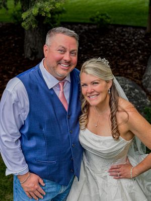 A smiling bride in a white gown and veil poses with a man in a blue vest and pink tie outdoors.