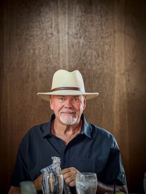 Smiling older man with white goatee wearing a cream panama hat and dark shirt sitting at a restaurant table.