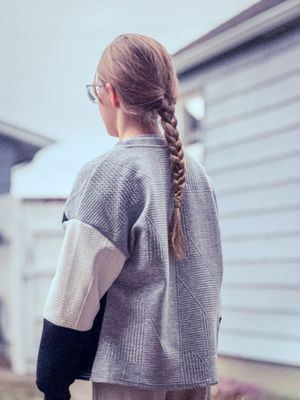 Young girl with long braided hair wearing gray plaid oversized jacket viewed from behind.