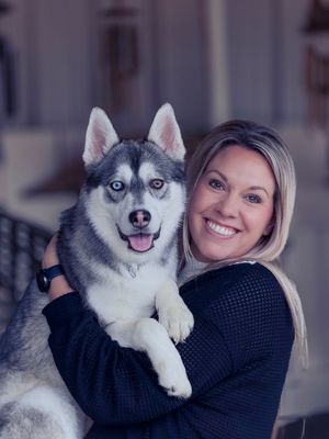Smiling blonde woman holding a gray and white Siberian Husky dog with heterochromia eyes outdoors.