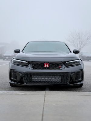 Front view of a dark gray Honda Civic Type R parked in a foggy parking lot, showcasing its aggressive front bumper and red H badge.