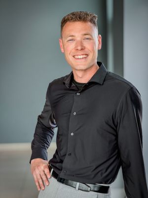 Smiling man in black button-down shirt and gray pants posing for professional headshot.