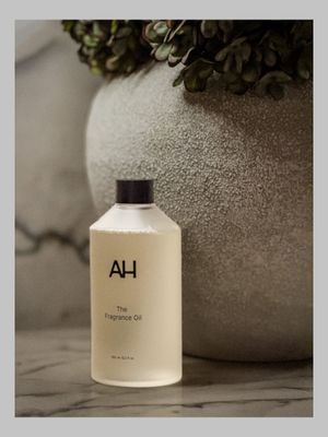 AH fragrance oil bottle in cream packaging on marble surface with stone vase and greenery.