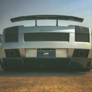 Silver Lamborghini Gallardo rear view with large spoiler and dual exhaust on open road.
