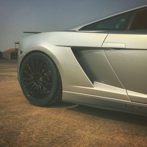 Silver Lamborghini Gallardo supercar with black wheels parked on open lot in warm light.