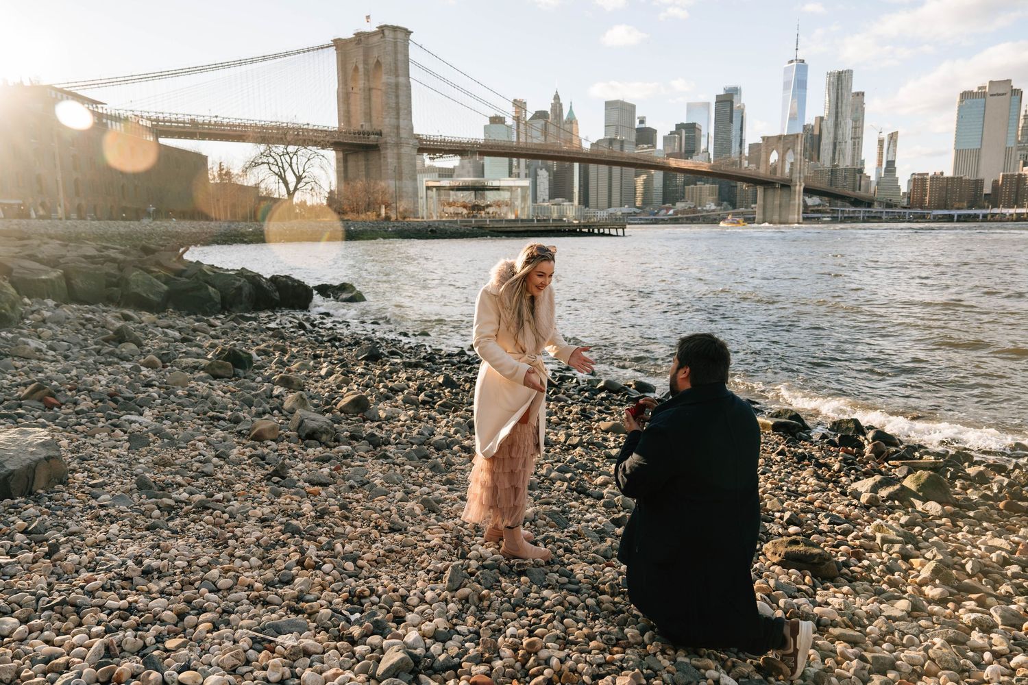 A woman in casual attire sits on the iconic Brooklyn Bridge during golden hour with the NYC skyline behind her.