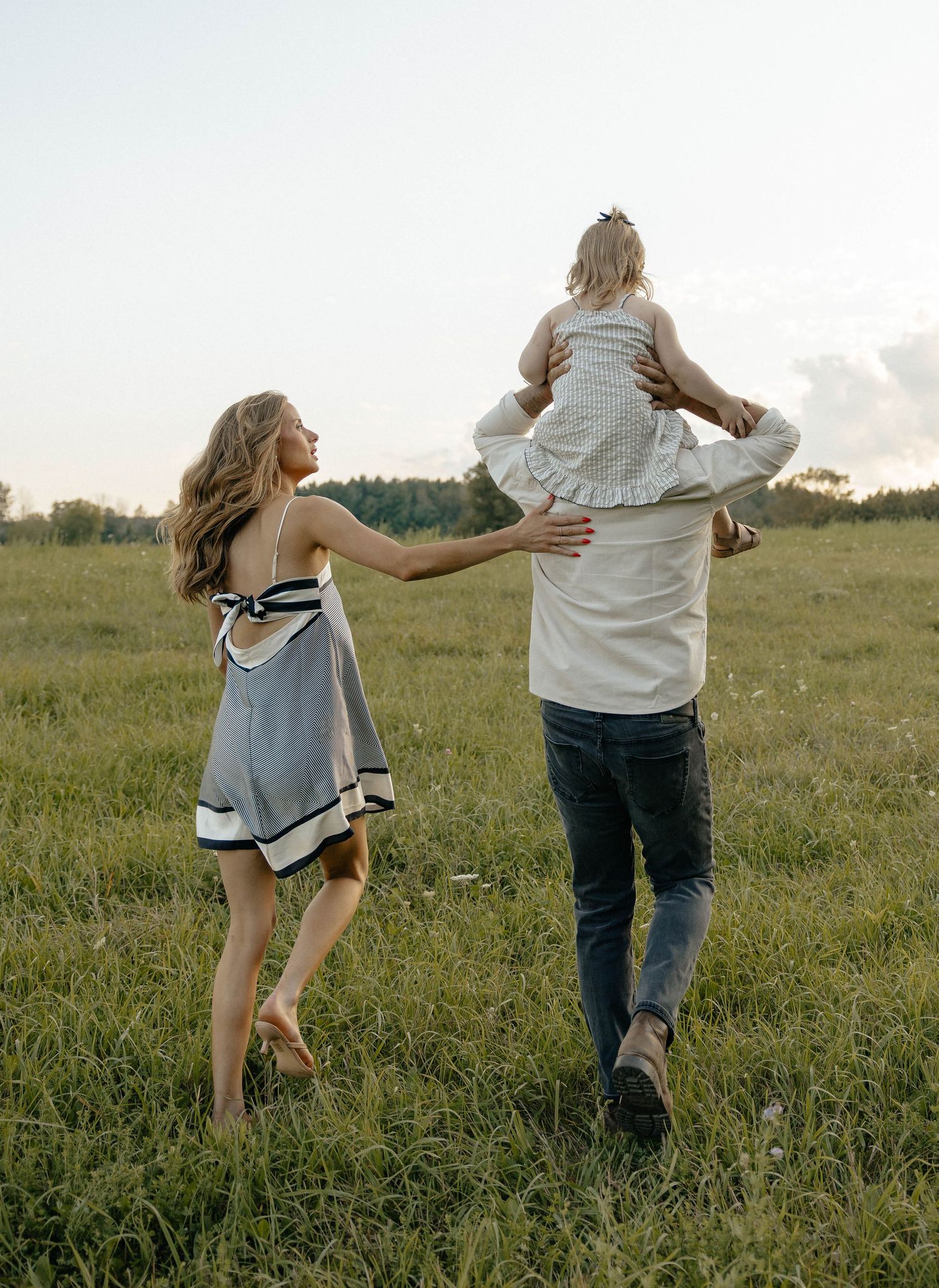 A family walks through a green field, father lifting young child overhead while mother reaches out joyfully.