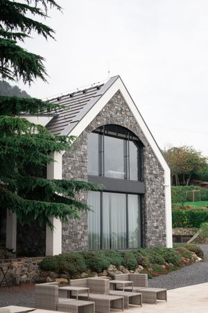 Modern stone and glass gabled wedding venue exterior with lush evergreen trees and outdoor seating.