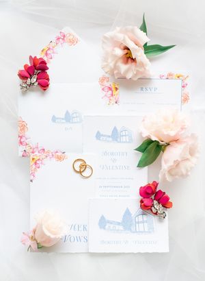 Flat lay of blue illustrated wedding stationery suite surrounded by pink peonies and red floral accents.