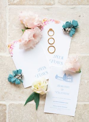 Wedding stationery with vow books, gold rings, and pink and teal flowers on stone.