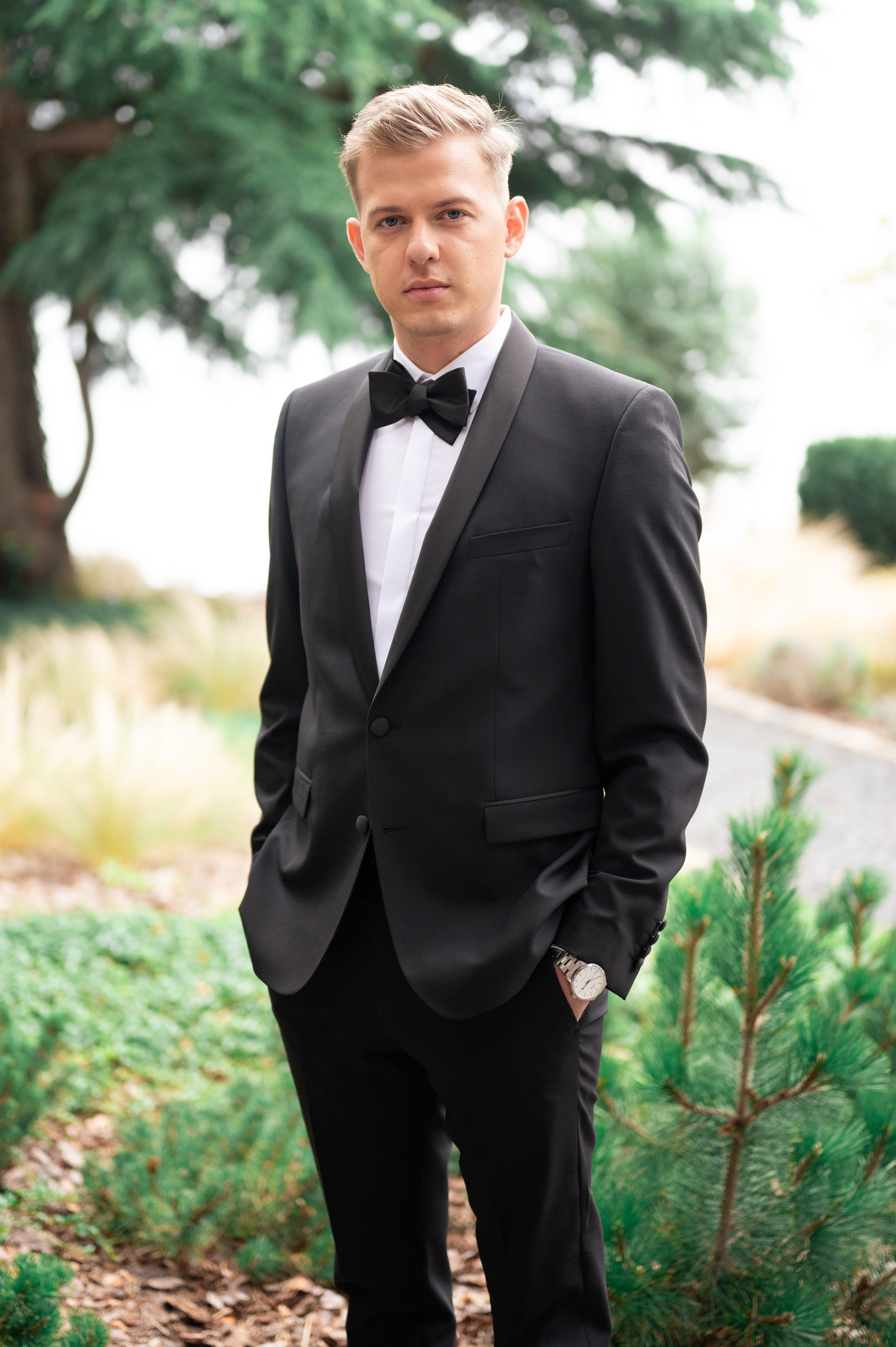 Handsome groom in black tuxedo with bow tie standing among pine shrubs outdoors.