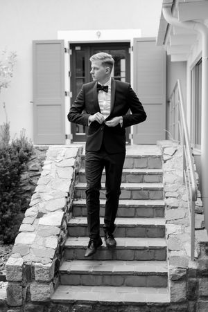 Groom in black tuxedo standing on stone steps outside building, black and white photo.