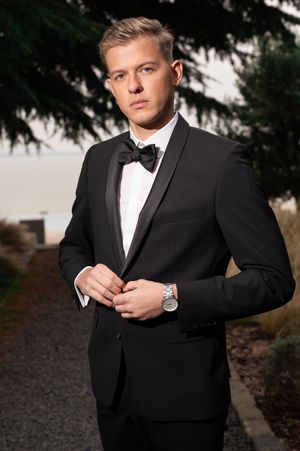 Young man in black tuxedo with bow tie and silver watch buttoning jacket outdoors.