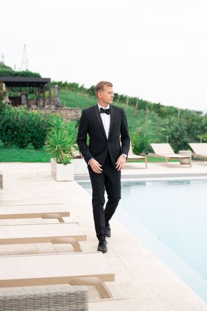 Elegant groom in black tuxedo walking beside luxury pool at outdoor wedding venue.