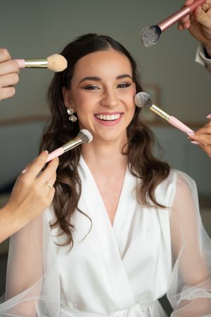 Laughing bride in white robe surrounded by makeup artists applying blush with pink brushes.