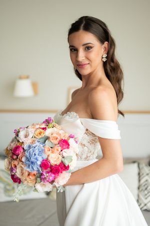 Bride in off-shoulder white gown holding colorful bouquet of roses and hydrangeas, smiling at camera.