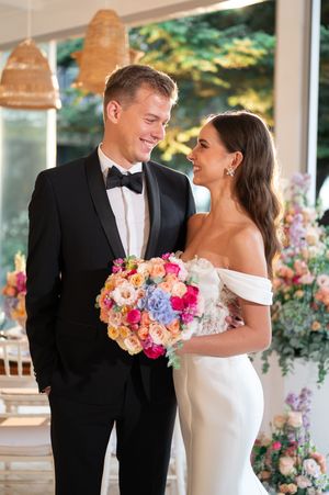 Smiling bride and groom in tuxedo holding vibrant colorful bouquet at bright modern wedding venue.