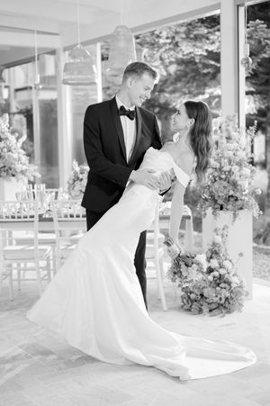 Black and white photo of groom in tuxedo dipping bride in fitted wedding gown at reception venue.