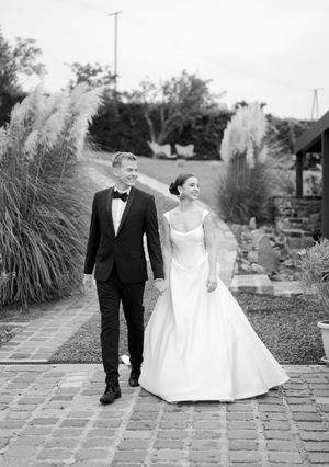 Black and white photo of bride and groom walking hand in hand on brick path through garden.