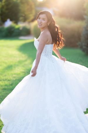 Smiling bride in a strapless white ball gown twirling in a sunlit garden, long dark hair flowing.
