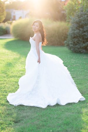 Bride in a flowing white strapless ball gown twirling on green grass with golden sunset light in background.