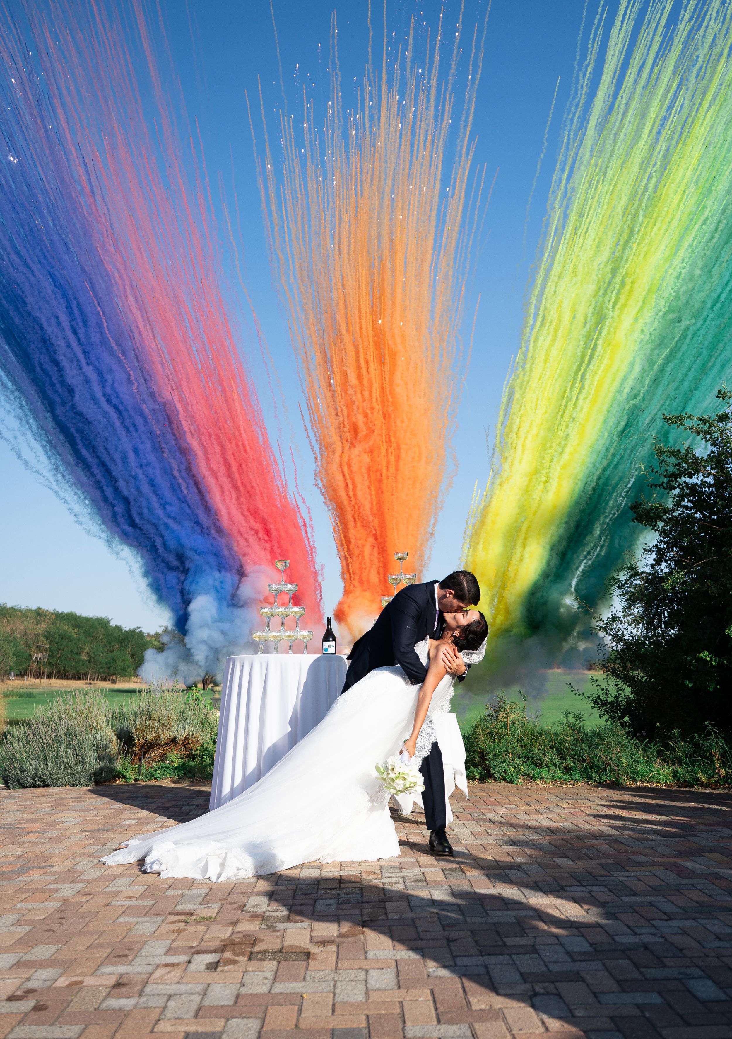 Bride and groom share a romantic dip kiss surrounded by vibrant rainbow colored smoke fountains at outdoor wedding.