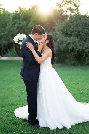 A bride and groom share a romantic moment outdoors, with golden sunlight glowing behind lush green trees.