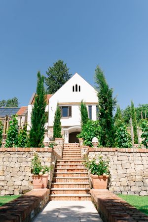 White wedding venue with stone steps, cypress trees, and terraced garden under blue sky.