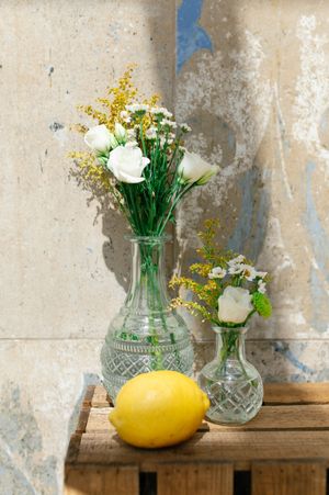 Two crystal vases with white roses and yellow wildflowers beside a lemon on a wooden crate.
