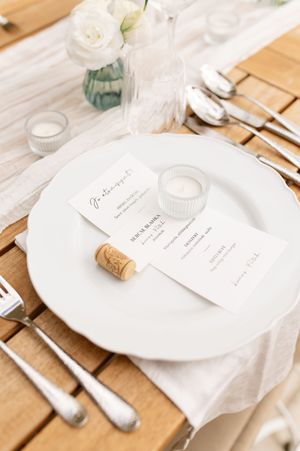 Elegant white wedding place setting with menu cards, wine cork, and floral centerpiece on wooden table.
