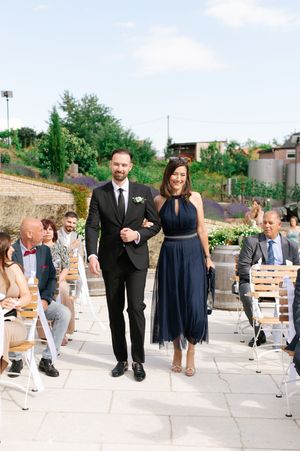 Groom in black suit escorts bridesmaid in navy dress down outdoor wedding ceremony aisle.