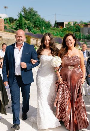 Bride walks down aisle escorted by parents in elegant outdoor wedding ceremony setting.
