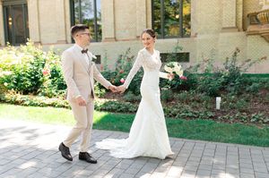 Bride in fitted lace gown and groom in beige suit hold hands while walking past a classic stone building outdoors.