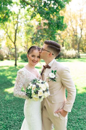 Groom in beige suit kisses bride's head as she smiles holding white floral bouquet in park.