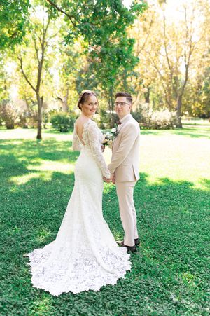 Bride in lace gown with train and groom in beige suit hold hands in lush green park.