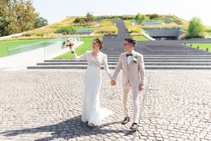 Bride and groom in beige suit walk hand in hand across cobblestone plaza with modern green hillside architecture behind them.
