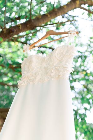 Elegant lace and floral applique wedding dress hanging from tree branch on wooden hanger.