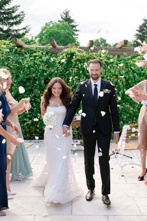 Happy couple walks through flower petal shower during outdoor wedding ceremony recessional with guests.