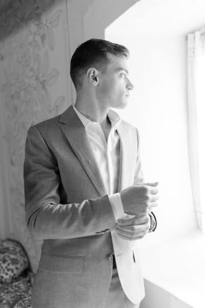 Groom in gray suit adjusts cufflinks while gazing out window in black and white portrait.