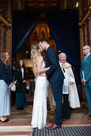 Newlyweds share a kiss at church entrance with priest and guests looking on joyfully.