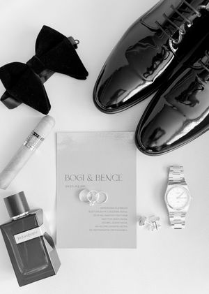 Black and white flat lay of groom wedding accessories including bow tie, patent shoes, rings, watch, and Bogi & Bence invitation.